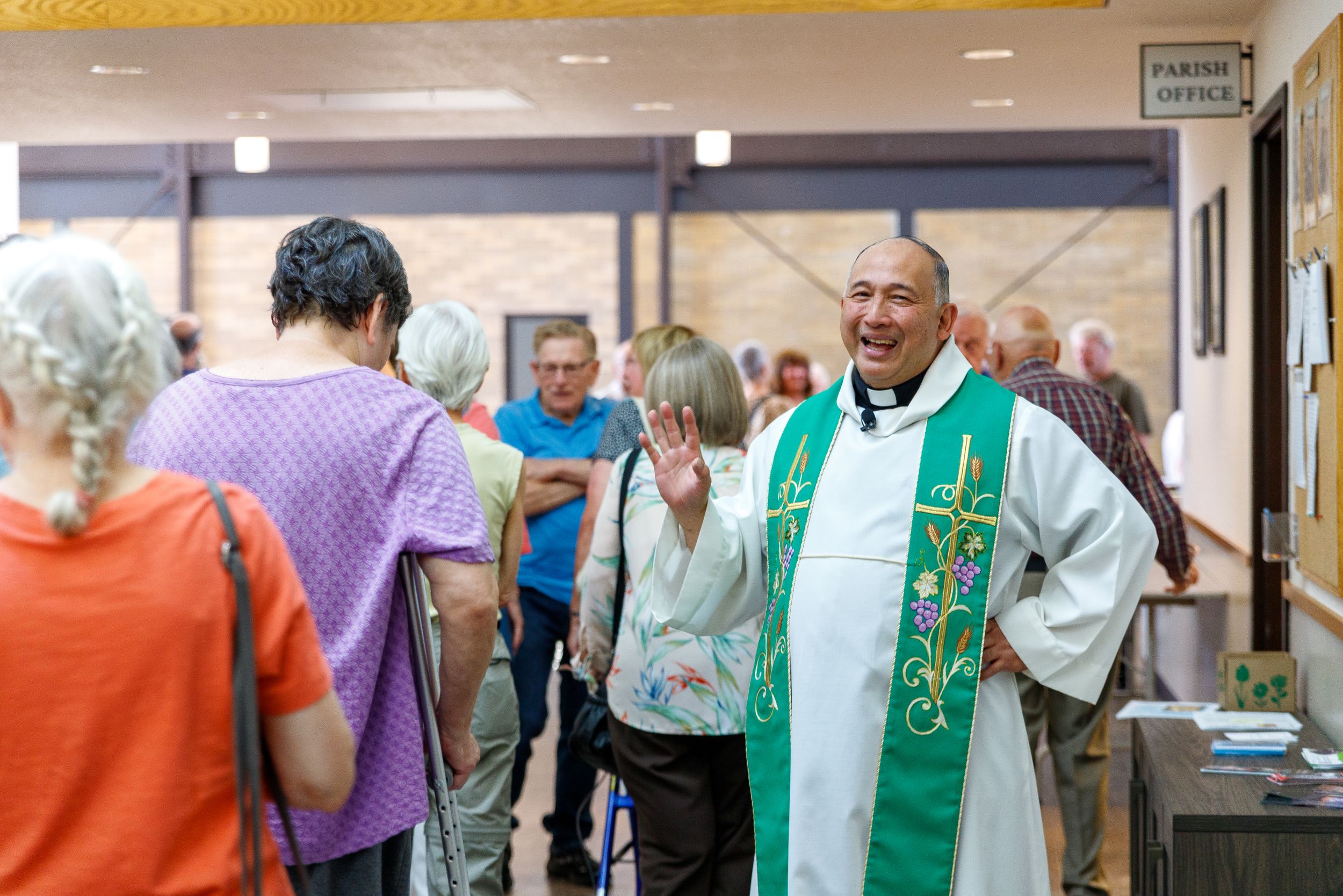 A priest in green and white vestments mingles with church members in a crowded hallway.