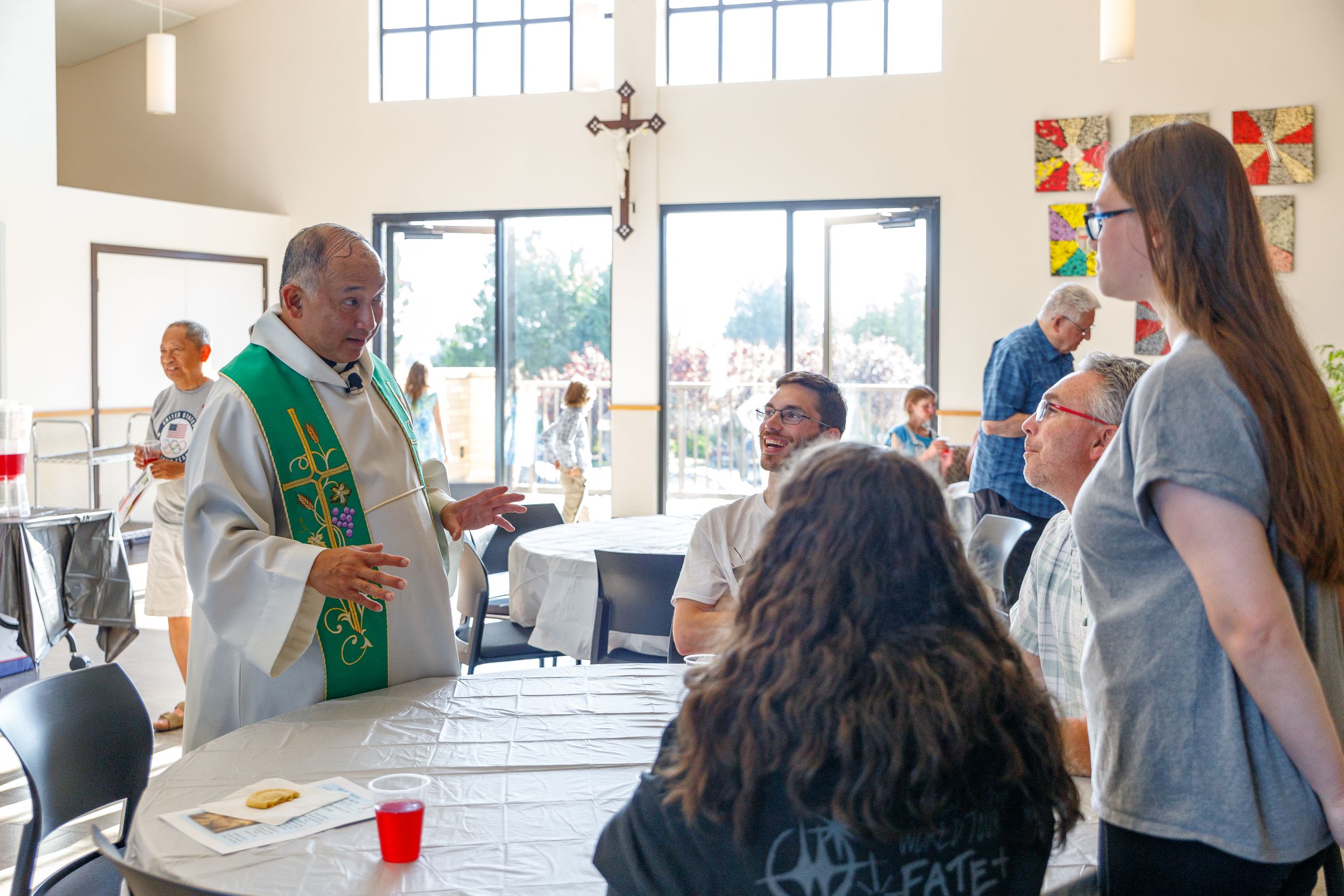 A minister in vestments converses with congregants around tables at a church social gathering.