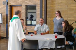 A minister in white robes speaks with parishioners seated at a table during a church event.