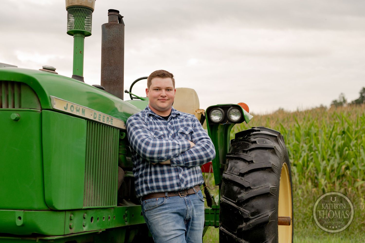 A person in plaid shirt poses against the wheel of a classic green John Deere tractor.