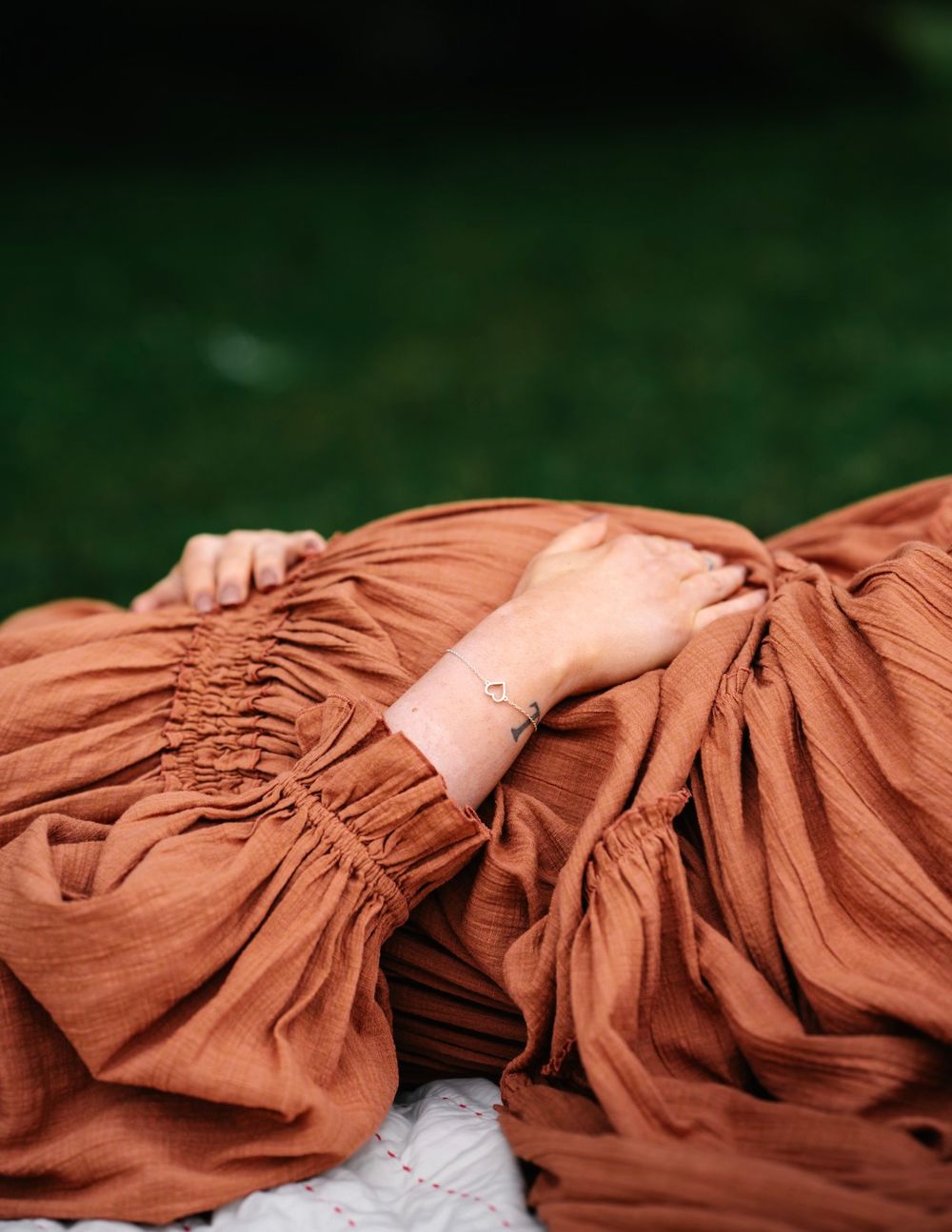 Woman in rust-colored linen dress with smocked sleeves lying on white blanket outdoors, wearing delicate bracelet.