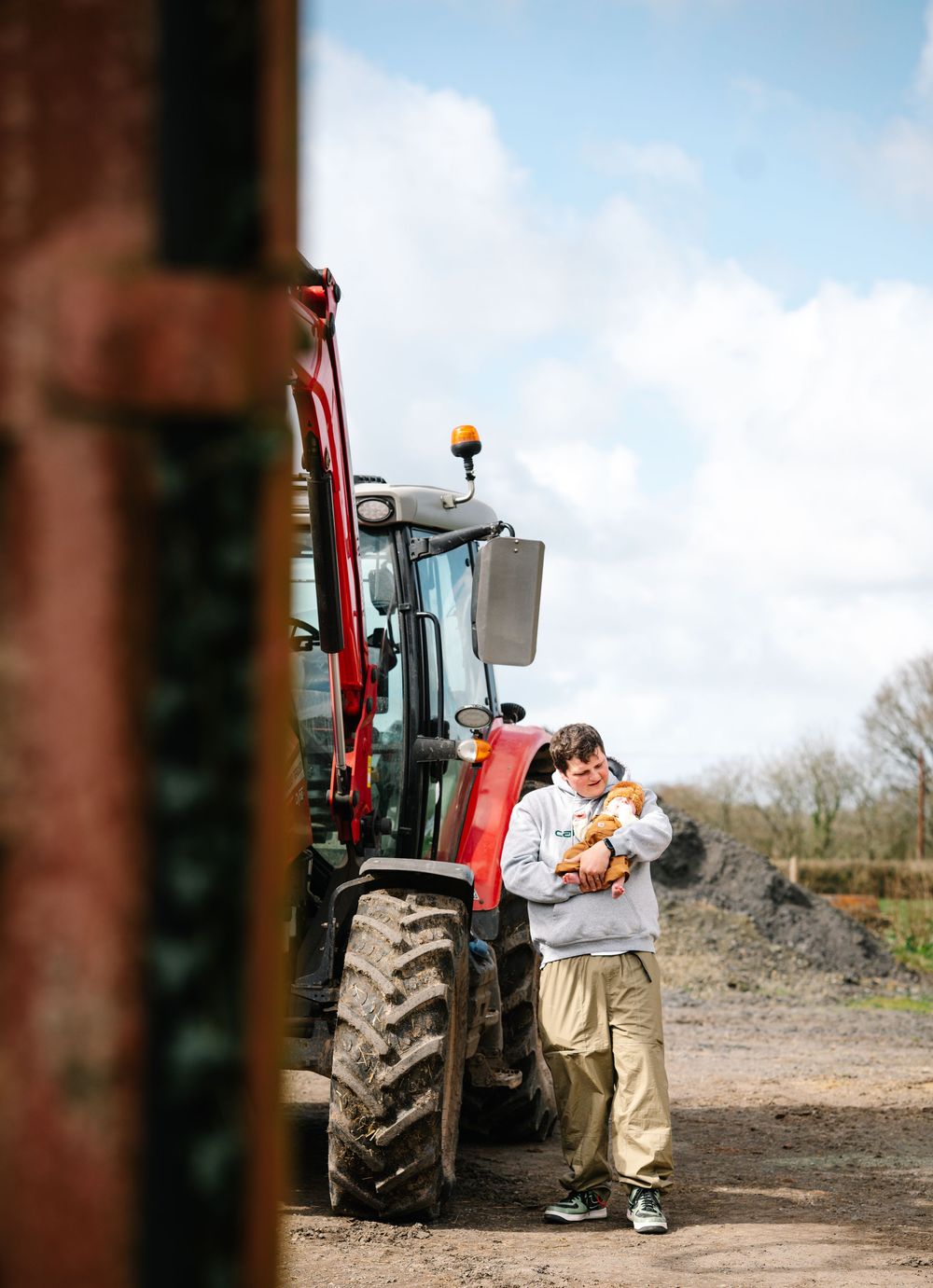 A young boy holds a small animal beside a red tractor on a farm under a partly cloudy sky.