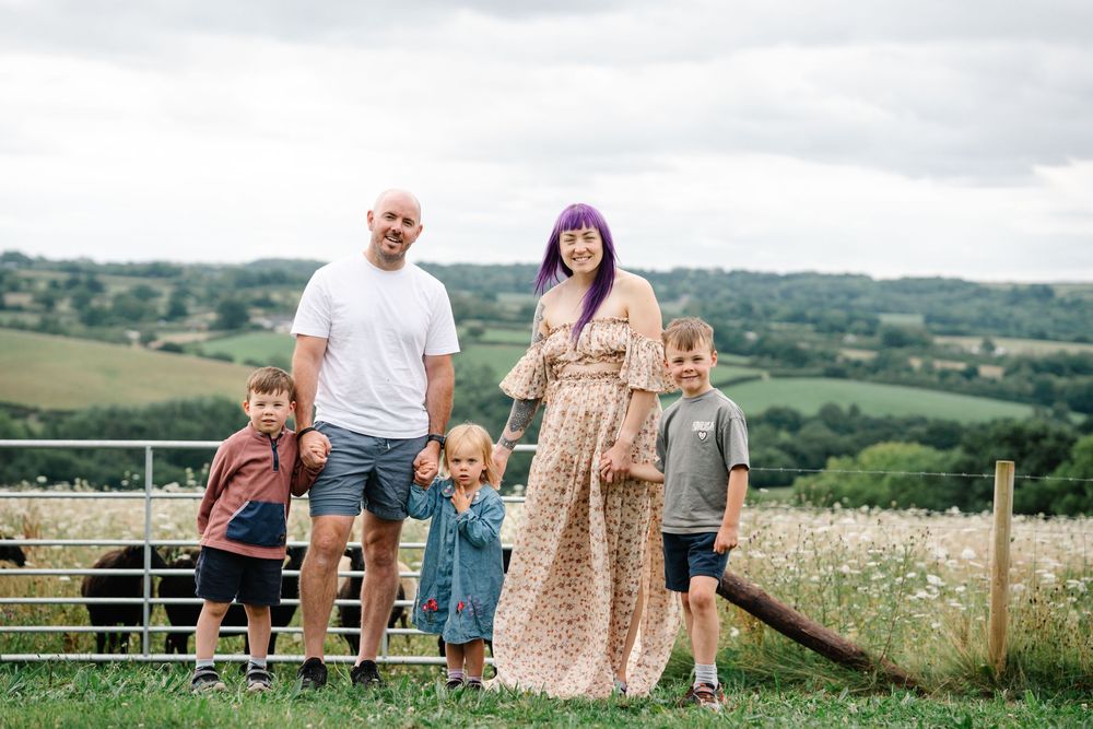 Family of five posing outdoors on a green hillside with scenic countryside views on a cloudy day.