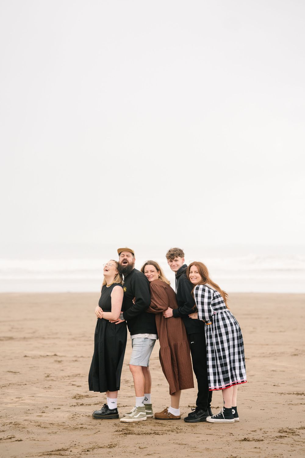 Five friends pose playfully on a sandy beach, laughing and leaning together under an overcast sky.