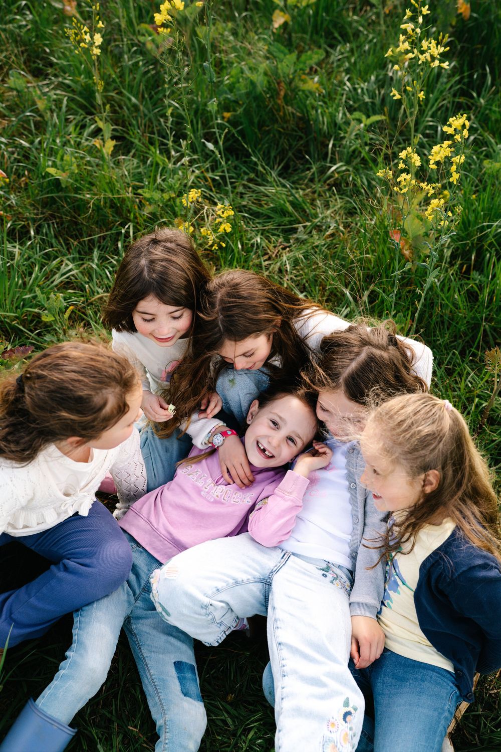 Group of young girls hugging and laughing together while sitting in a green meadow with yellow wildflowers.