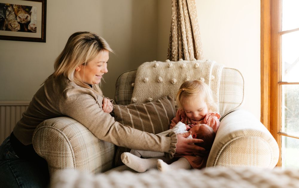 A smiling mother watches her toddler interact with a newborn baby in a cozy armchair by a sunlit window.