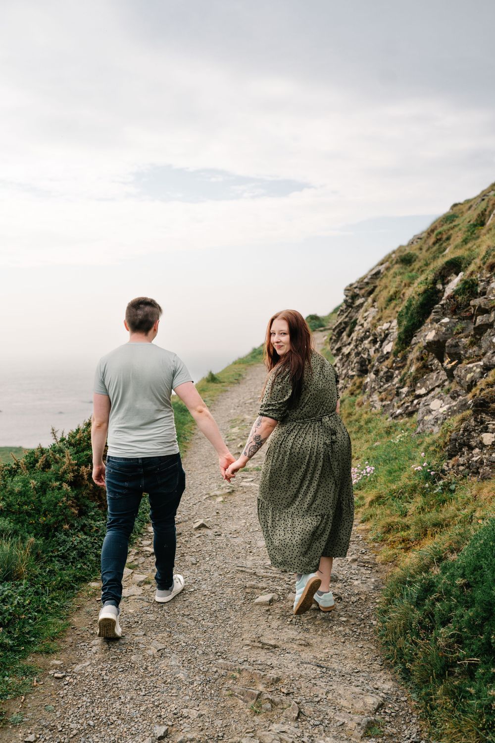 A couple holds hands walking a coastal cliff path, woman in green dress looks back smiling, rocky hillside backdrop.