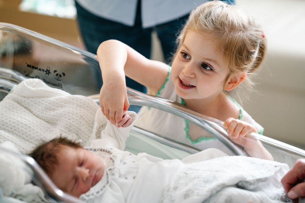 A young girl peers over the edge of a hospital bassinet at a newborn baby.