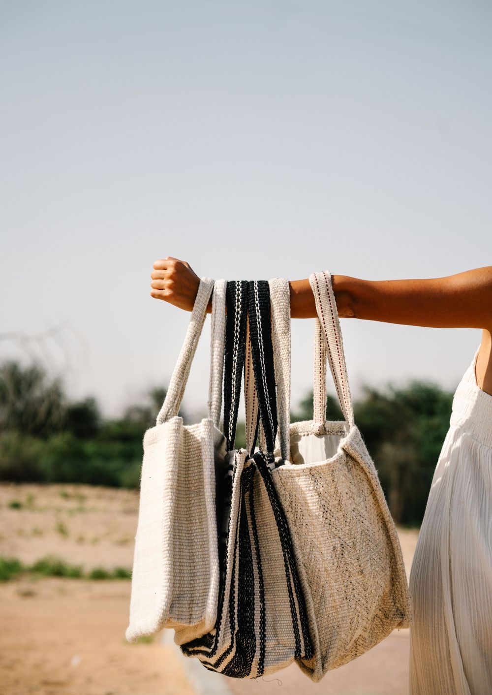 An arm holds multiple woven beach bags against a desert landscape.