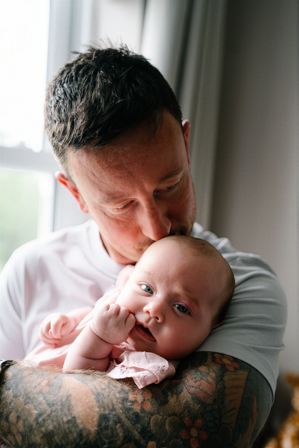 A tattooed father tenderly kisses his newborn baby dressed in pink near a bright window.