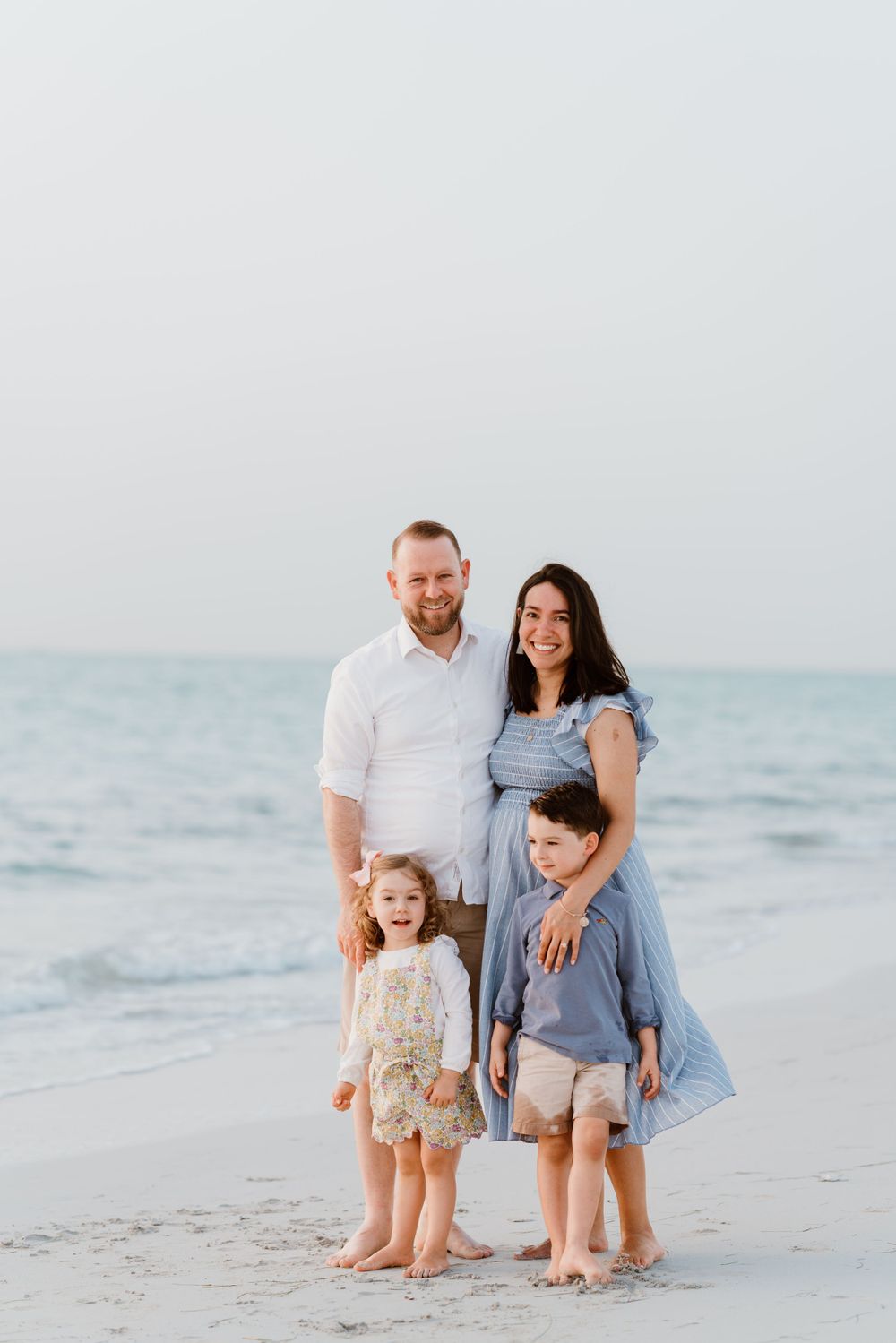 Family of four poses together on a beach during sunset in coordinated blue and white outfits.