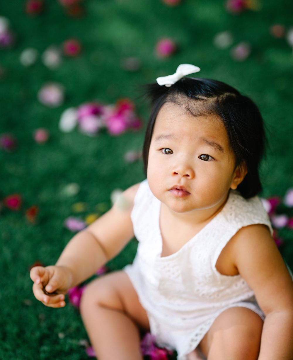 Baby in white outfit sitting on grass with colorful flowers in background.