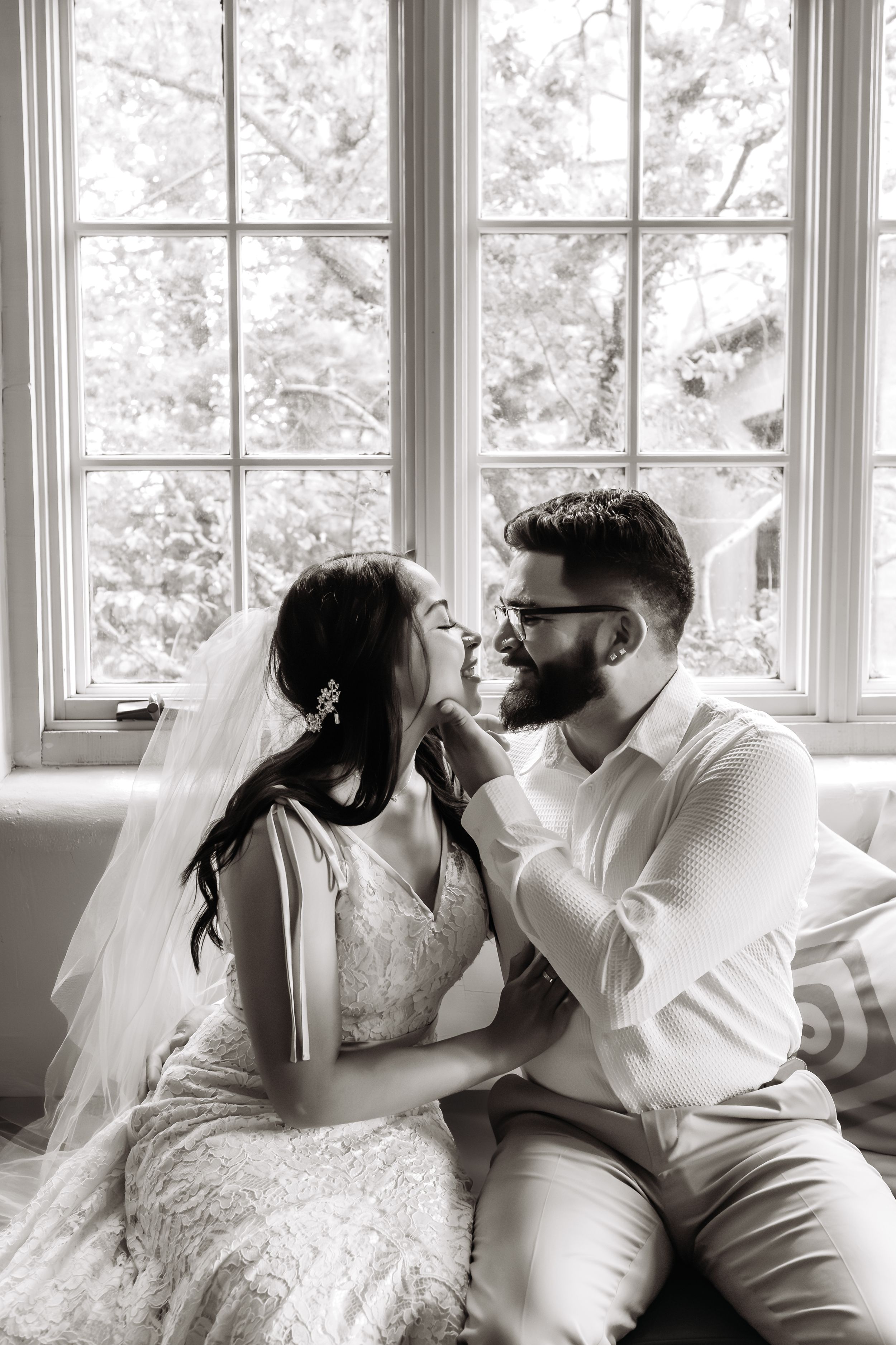 A romantic black and white photo of a couple in wedding attire sharing an intimate moment by a bright window.