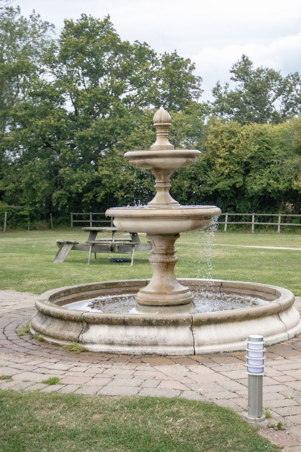 Stone fountain with circular basin sits in landscaped garden area with benches.