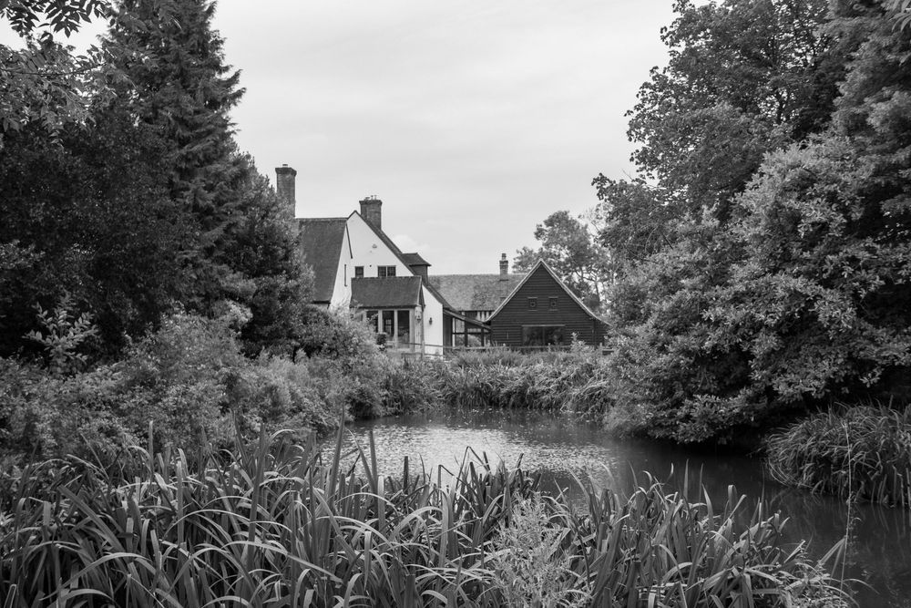 Black and white landscape of traditional houses reflected in a tranquil pond surrounded by reeds and trees.