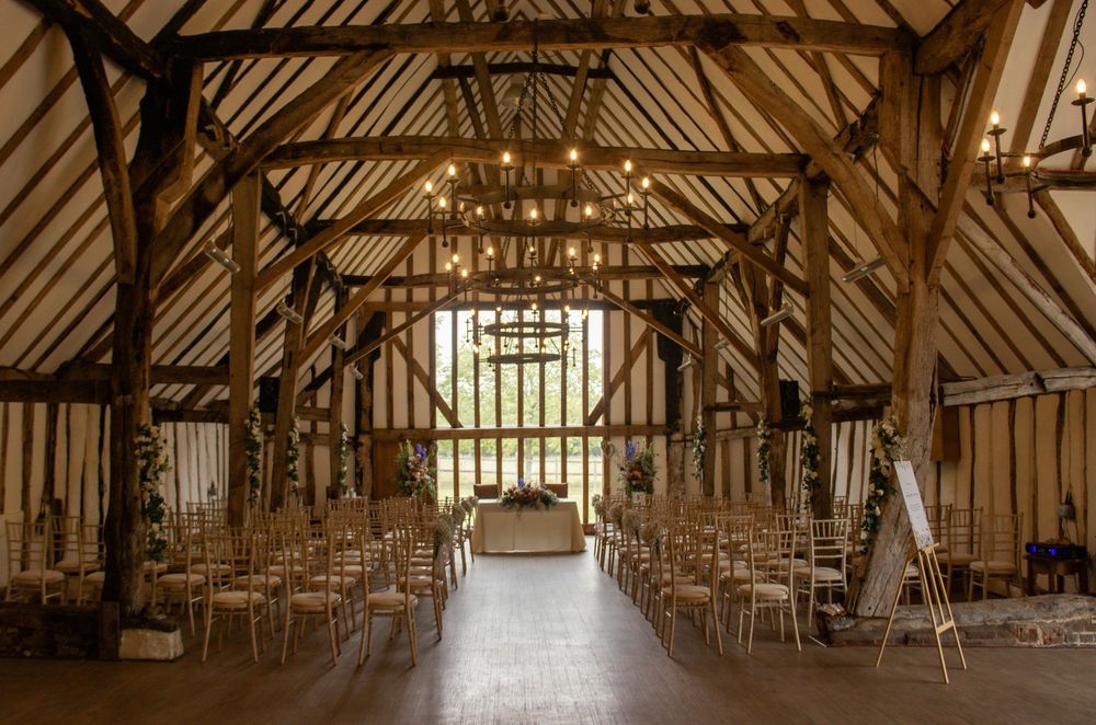 Interior of a traditional timber-framed barn with rows of white chairs set up for a wedding ceremony.