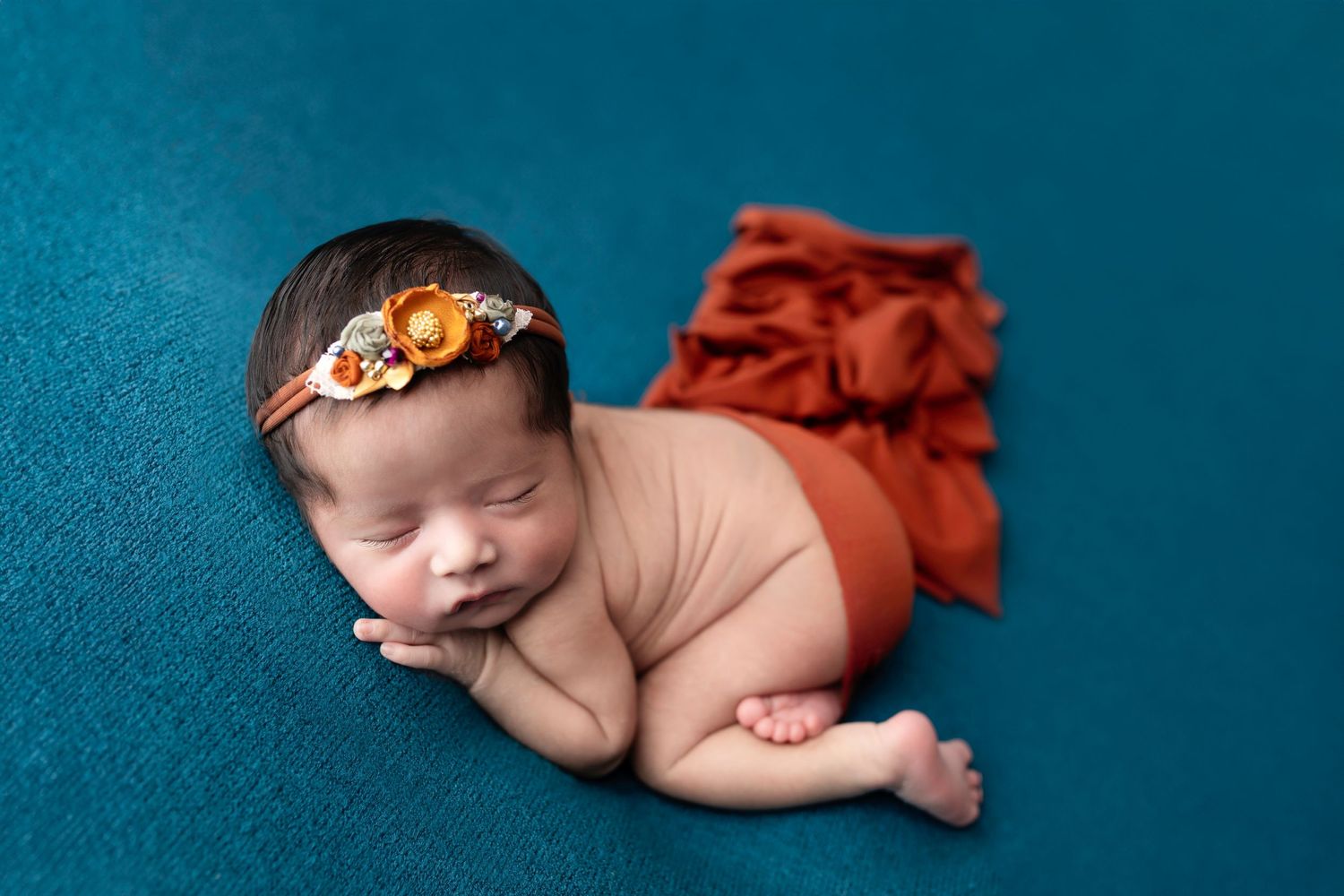 Sleeping newborn on turquoise background wearing orange tutu and floral headband poses for professional photo.
