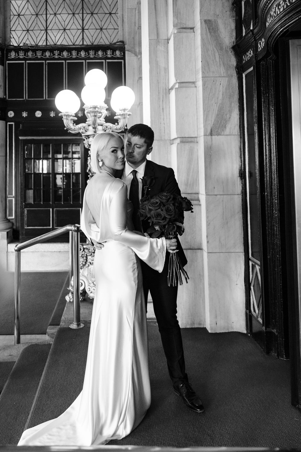 A black and white wedding photo series showing a couple dancing in an elegant hallway with ornate architecture.