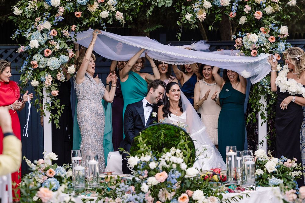 Wedding party celebrates under a floral arch with white fabric being held up during an outdoor ceremony.