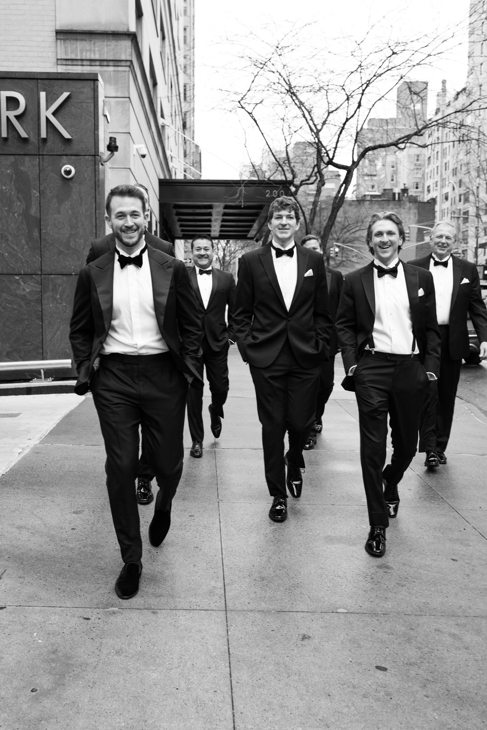 Groomsmen in black tuxedos walk confidently down a city street.