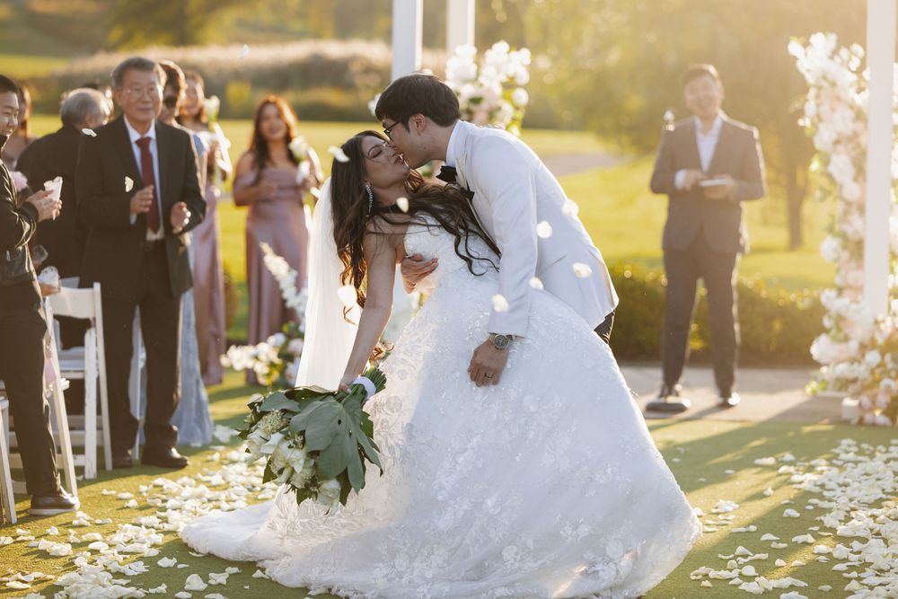 A couple in wedding attire shares a tender moment during their outdoor ceremony as guests look on in golden sunlight.