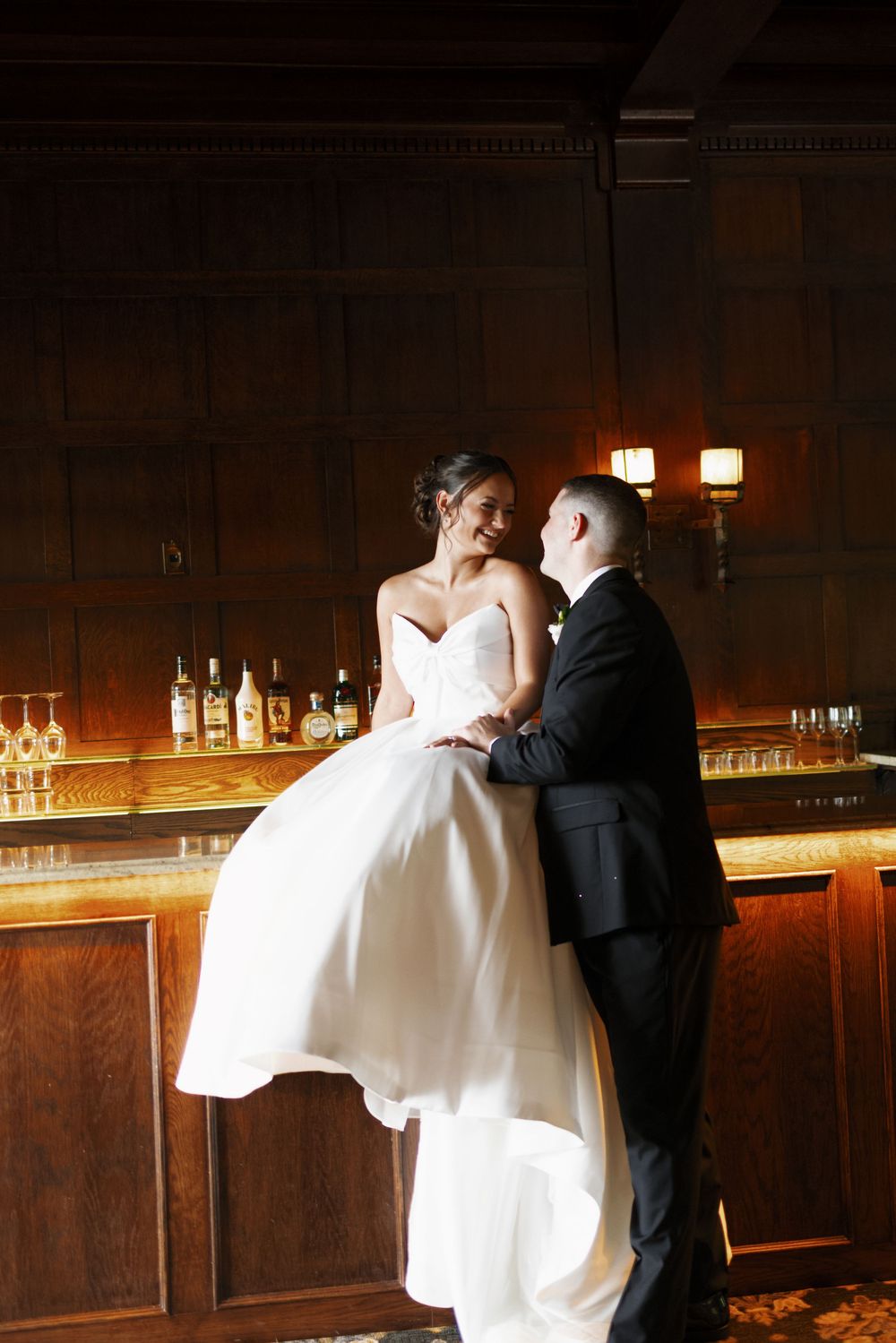 A couple in formal wedding attire share an intimate moment at a dimly lit wooden bar.