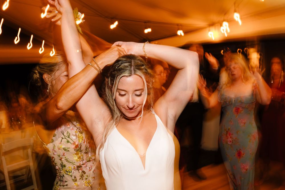 Wedding guests dance joyfully with raised arms on a warmly lit dance floor during an evening reception celebration.
