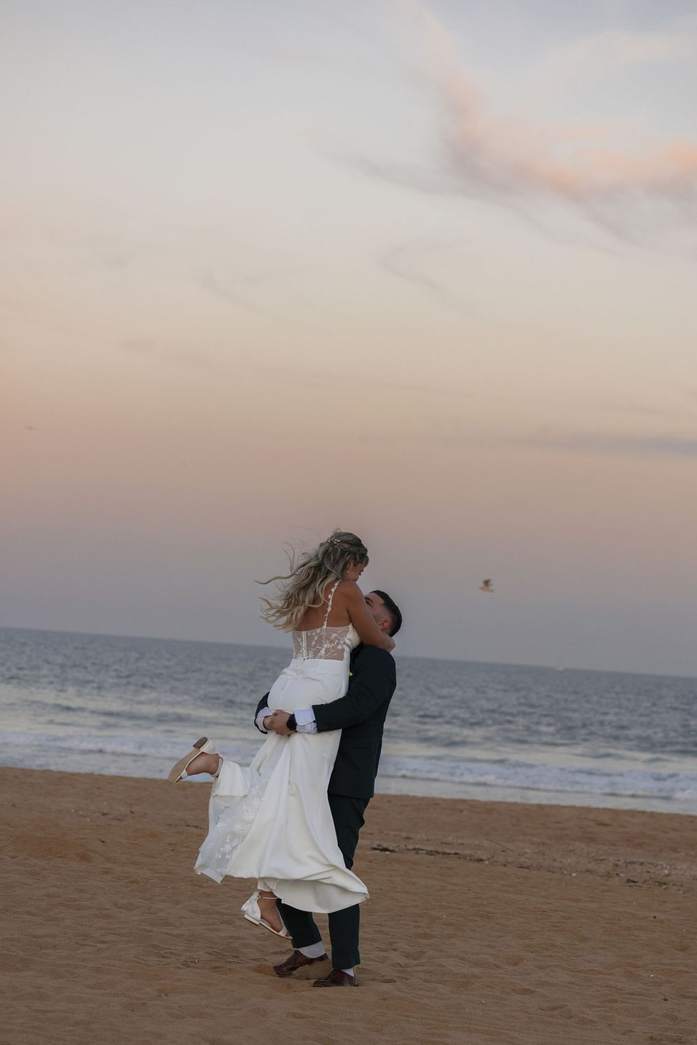 A couple in formal wedding attire dances and embraces on a sandy beach at sunset.