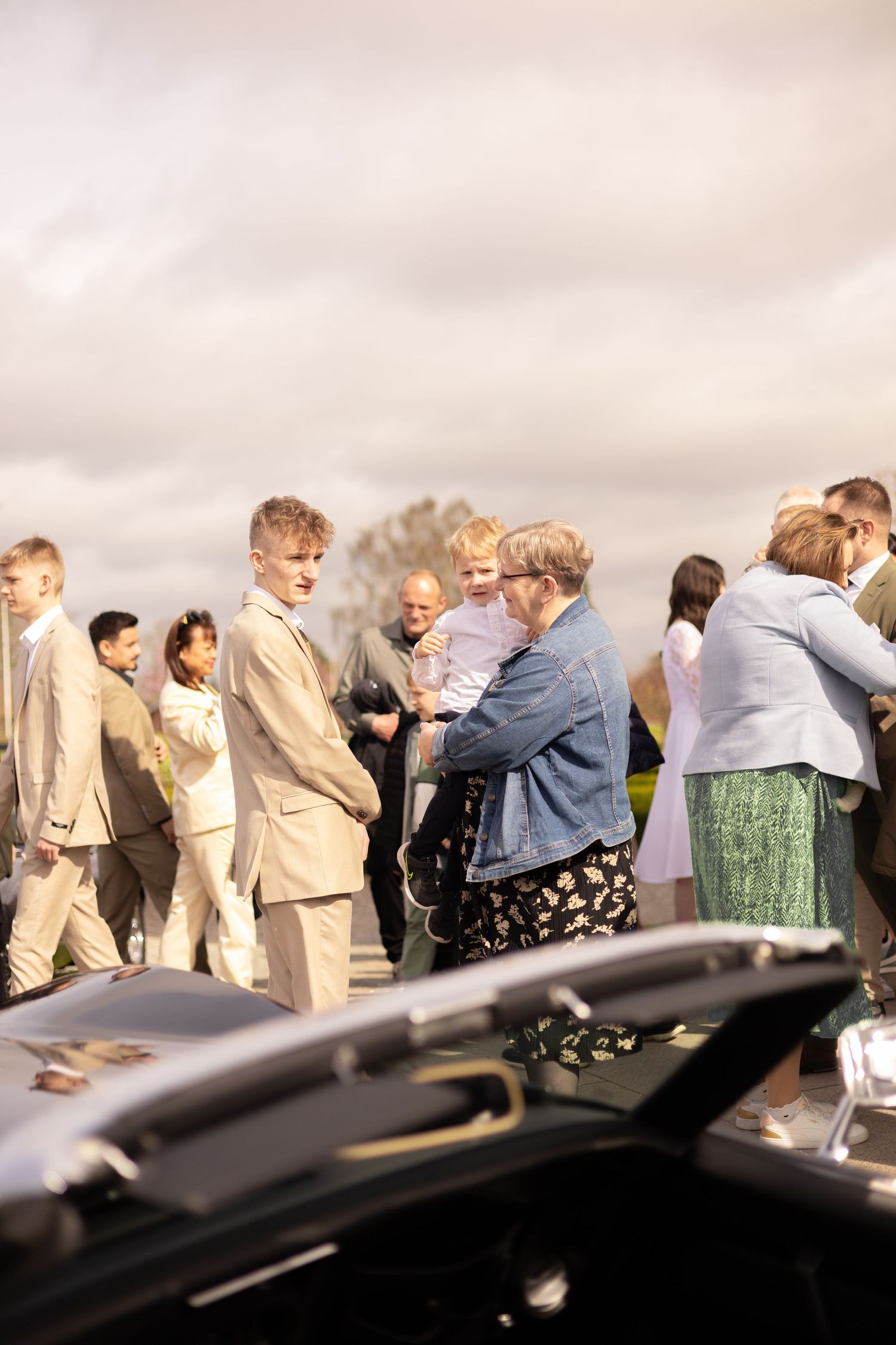 Outdoor gathering of formally dressed guests near a parked car.
