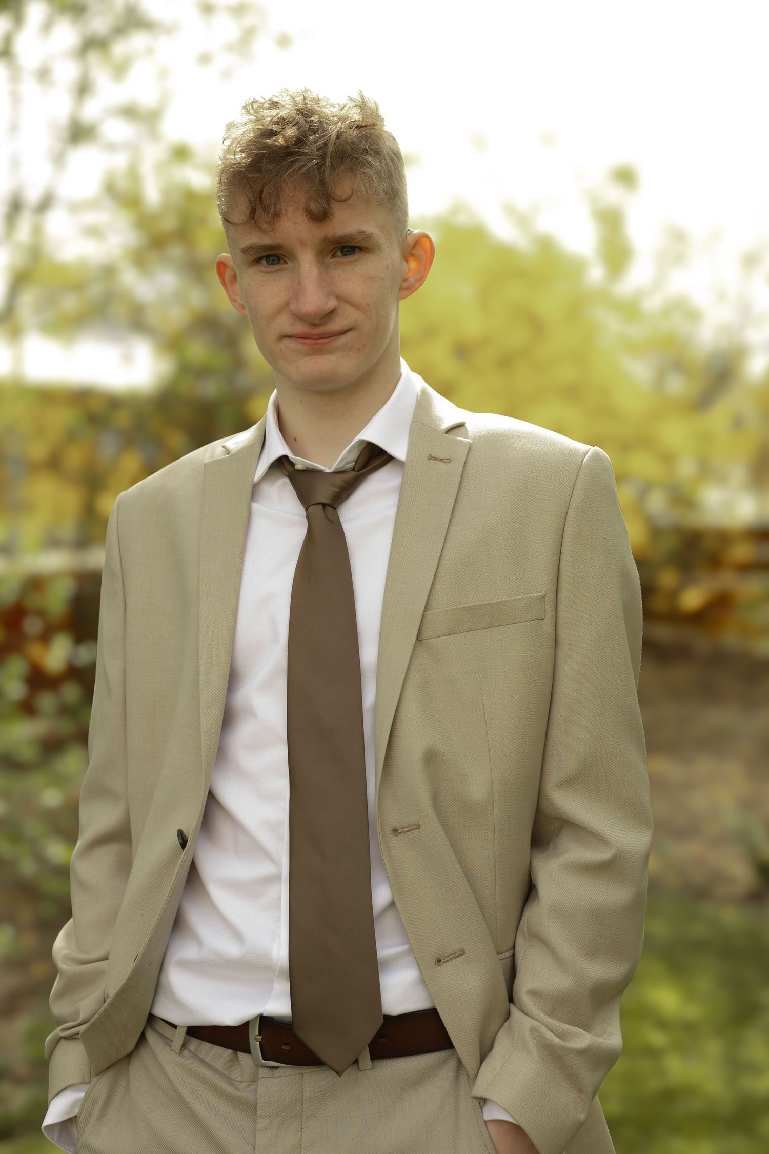 Young person in a tan suit and brown tie poses outdoors in natural lighting.