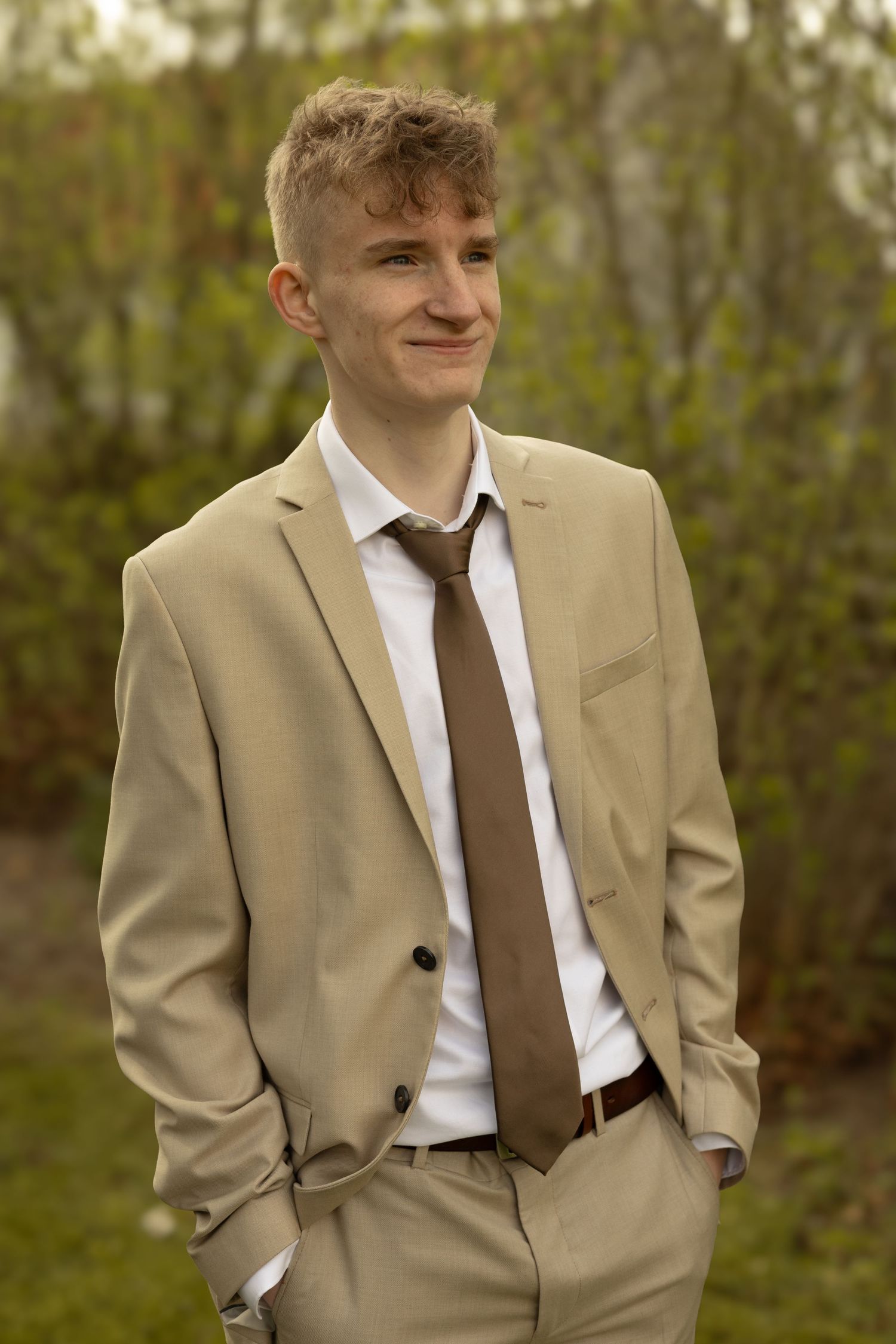 Person wearing beige suit with brown tie stands against blurred outdoor background.