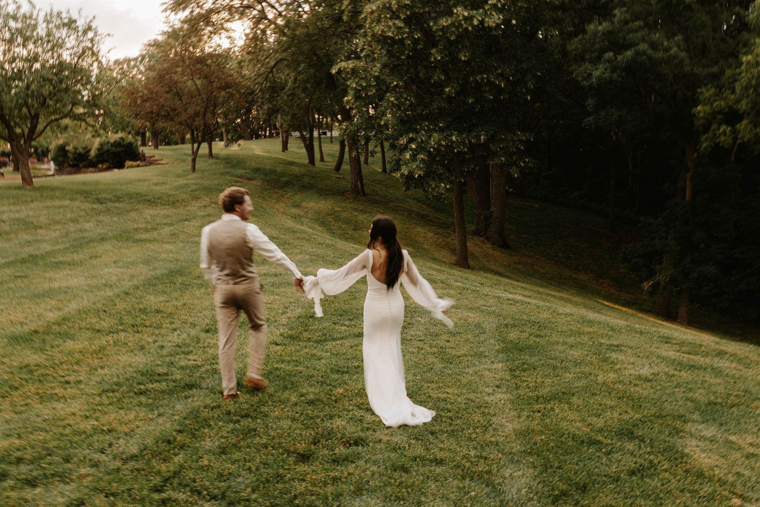 A couple holds hands while walking on a grassy hillside during golden hour.