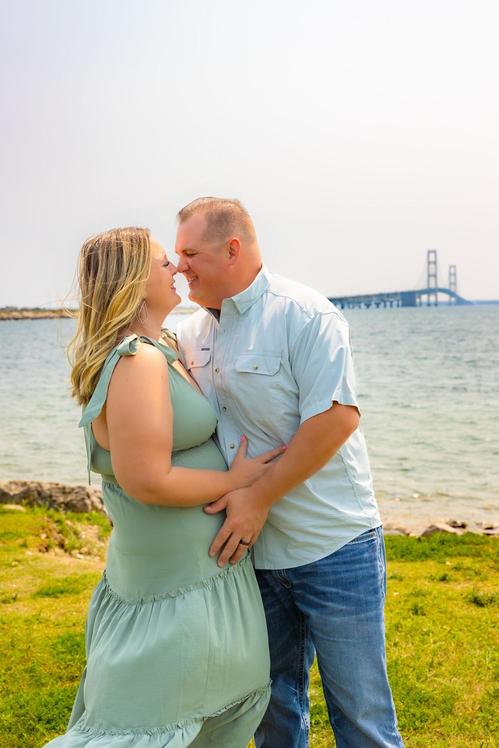 Romantic couple embraces by the Mackinac Bridge on a sunny summer day with water and grassy shore in view.