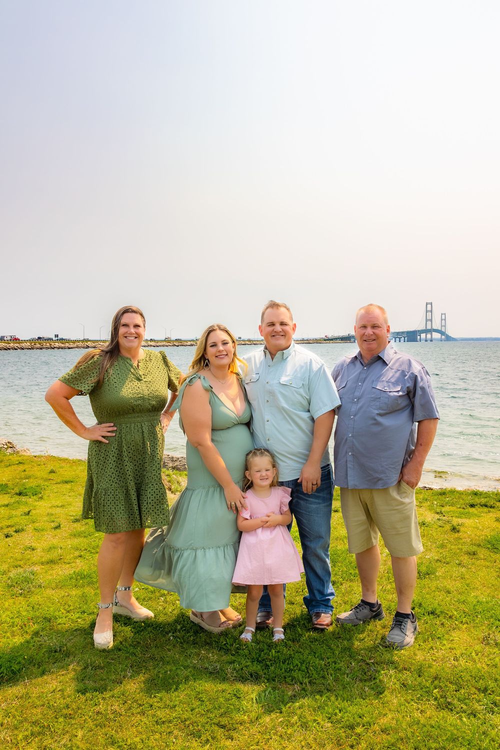 Family group poses together on grassy waterfront with Mackinac Bridge visible in background on summer day.