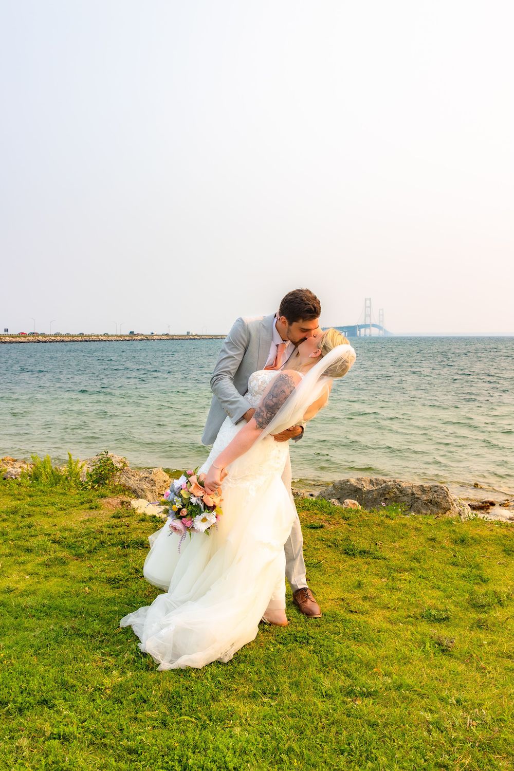 A romantic wedding couple dips in a dramatic pose against a coastal backdrop with green grass and ocean views.