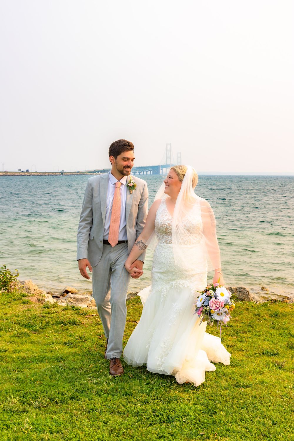 A couple in wedding attire walks along a scenic oceanfront lawn with blue water in the background.
