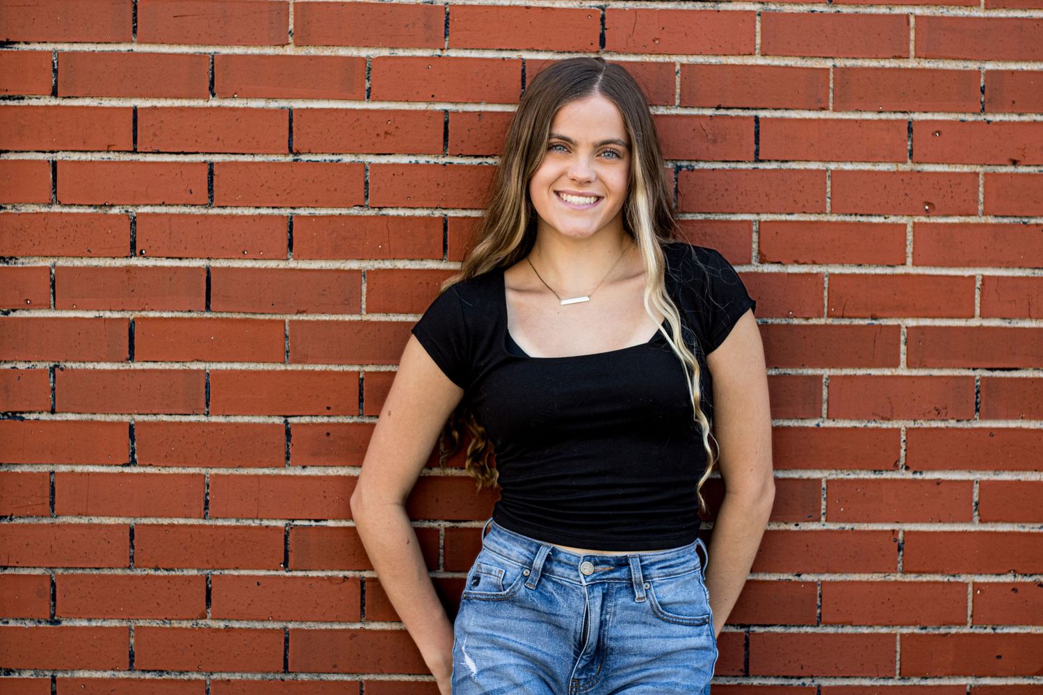 Young woman in black top and jeans smiling against a red brick wall outdoors.