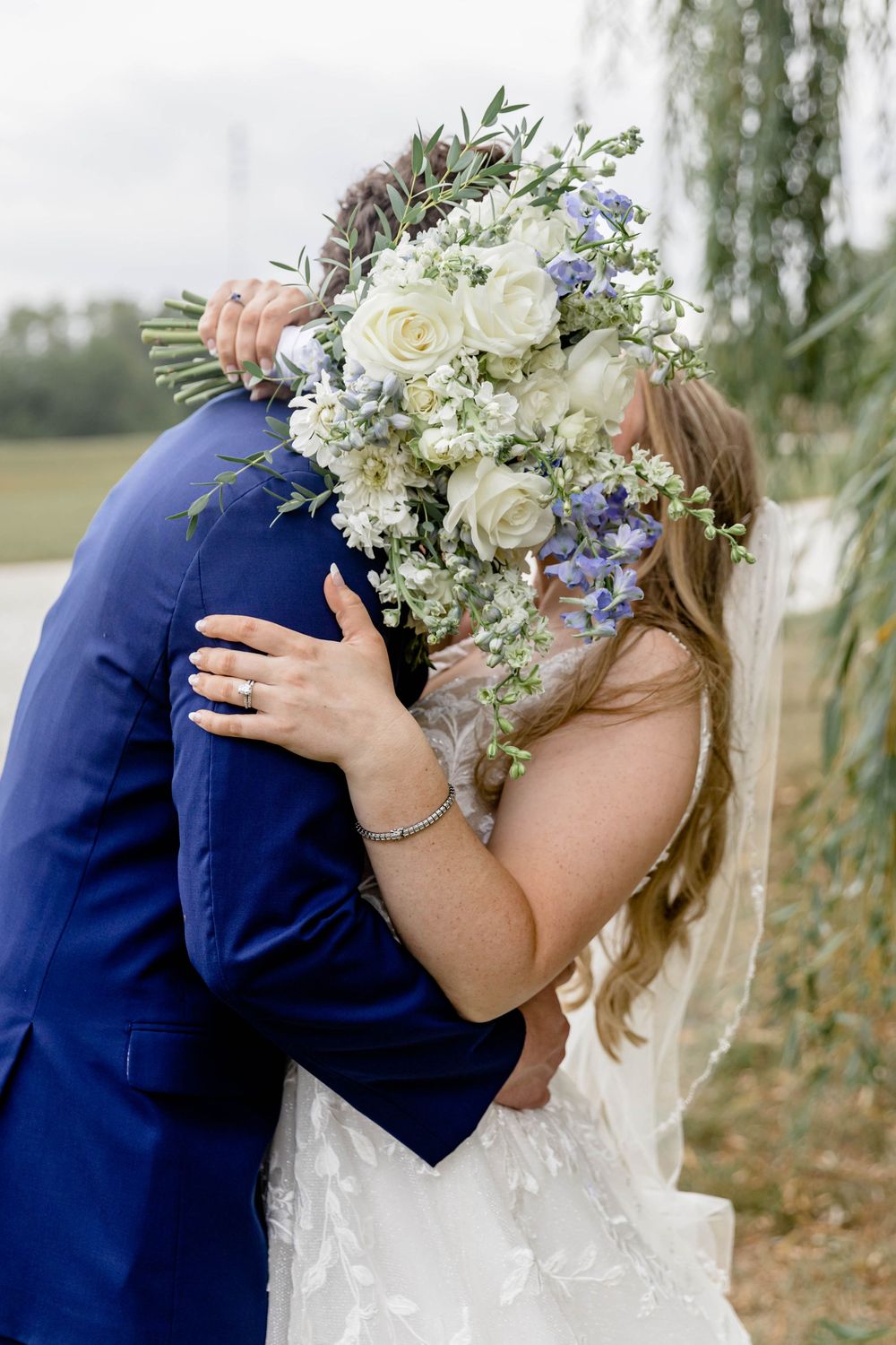 Wedding couple embracing with white and blue floral bouquet against natural backdrop.