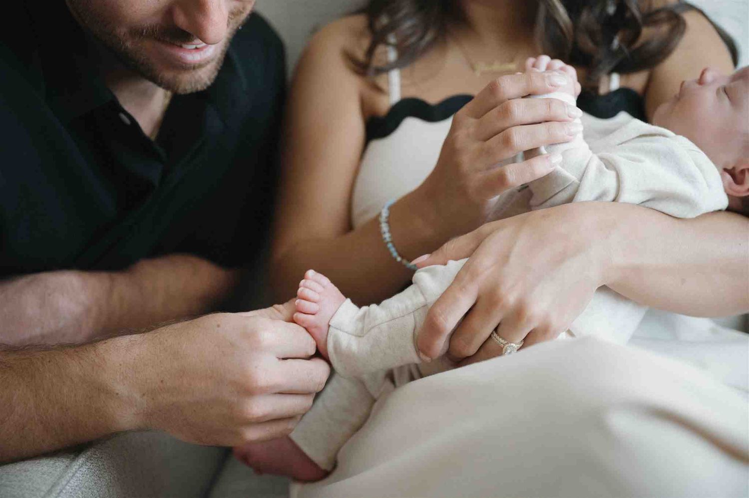 Close up of parents holding their newborn baby's tiny feet while sitting together on white bedding.
