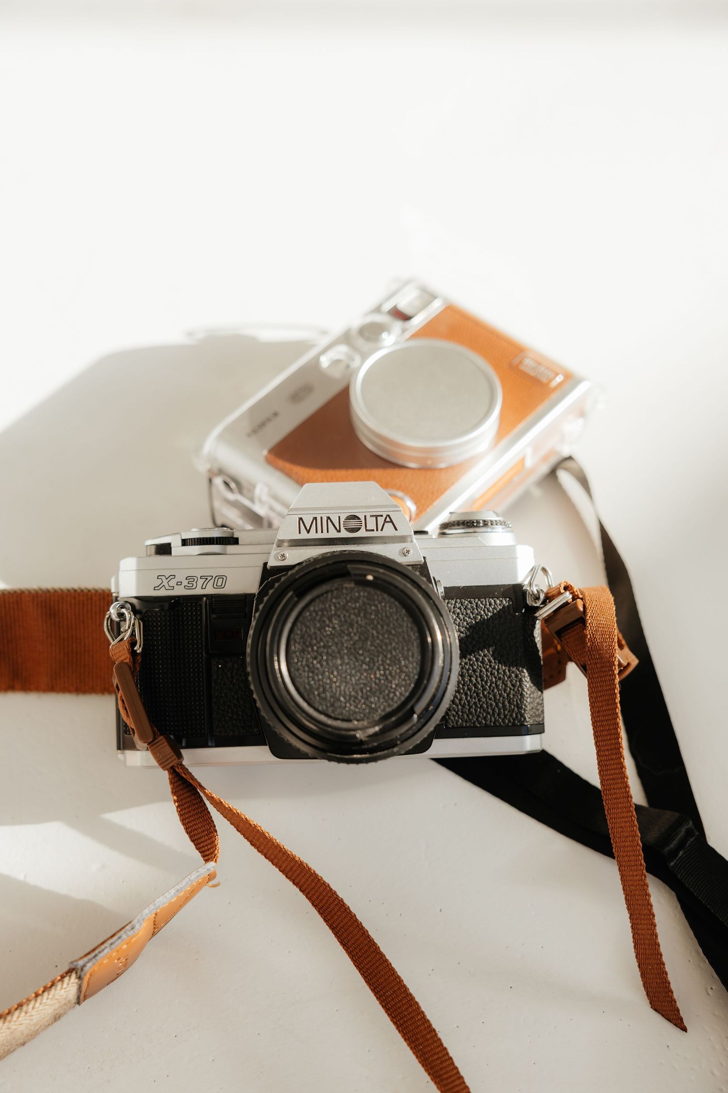 Vintage Minolta film camera with brown leather strap beside small instant camera on white surface.