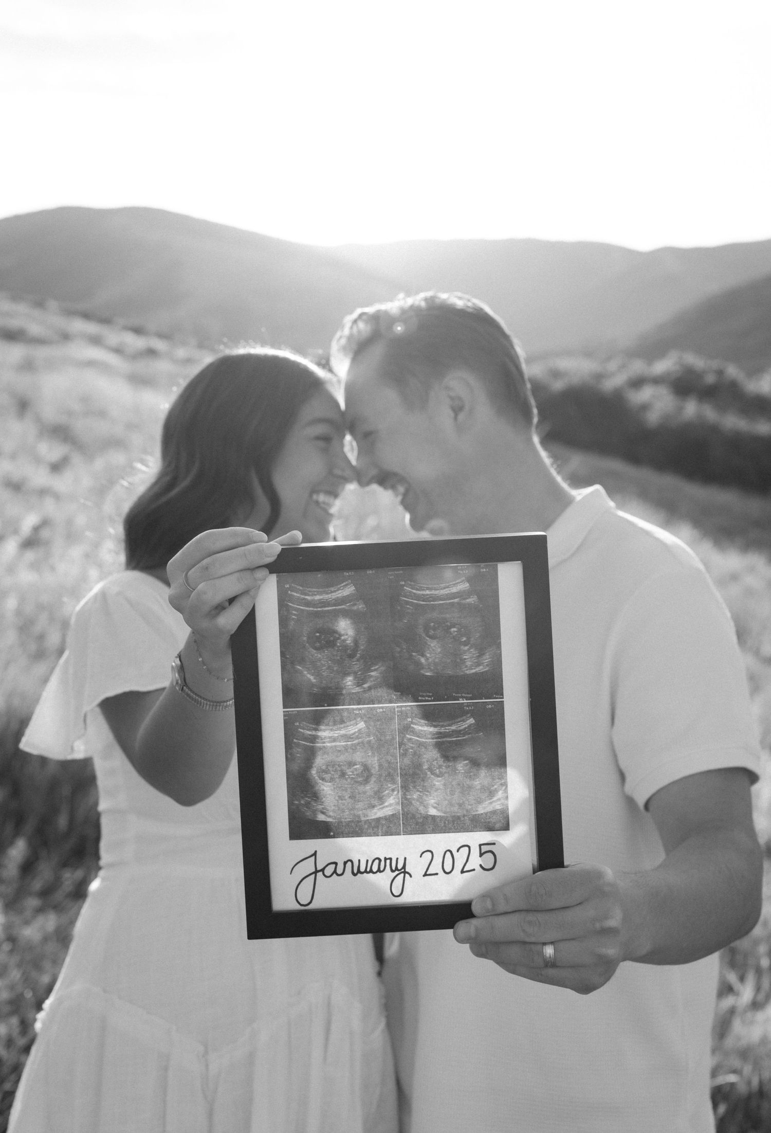 Black and white photo of a couple holding a framed ultrasound pregnancy announcement reading January 2025.