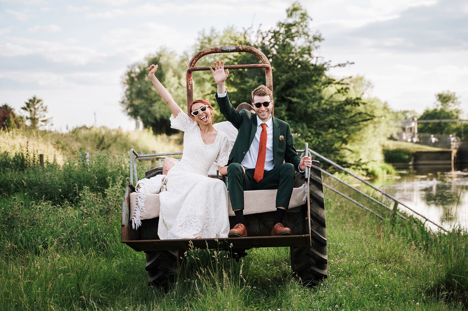 Wedding couple sitting playfully on a rustic cart by a canal with lush greenery.