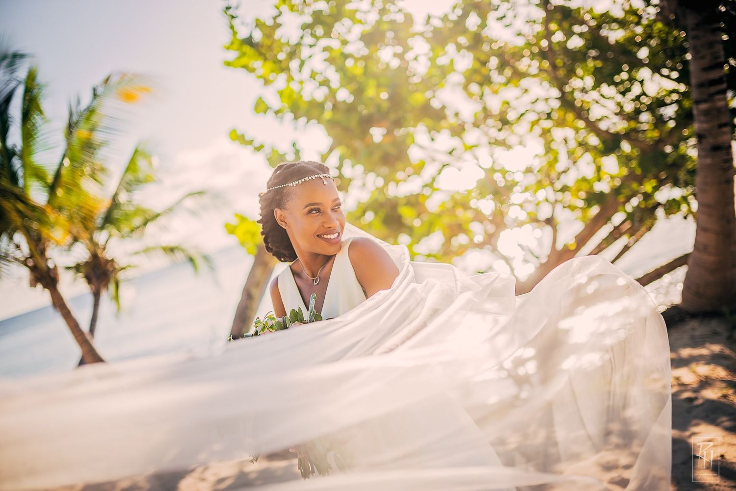 Joyful bride playing with flowing white veil against tropical beach backdrop.