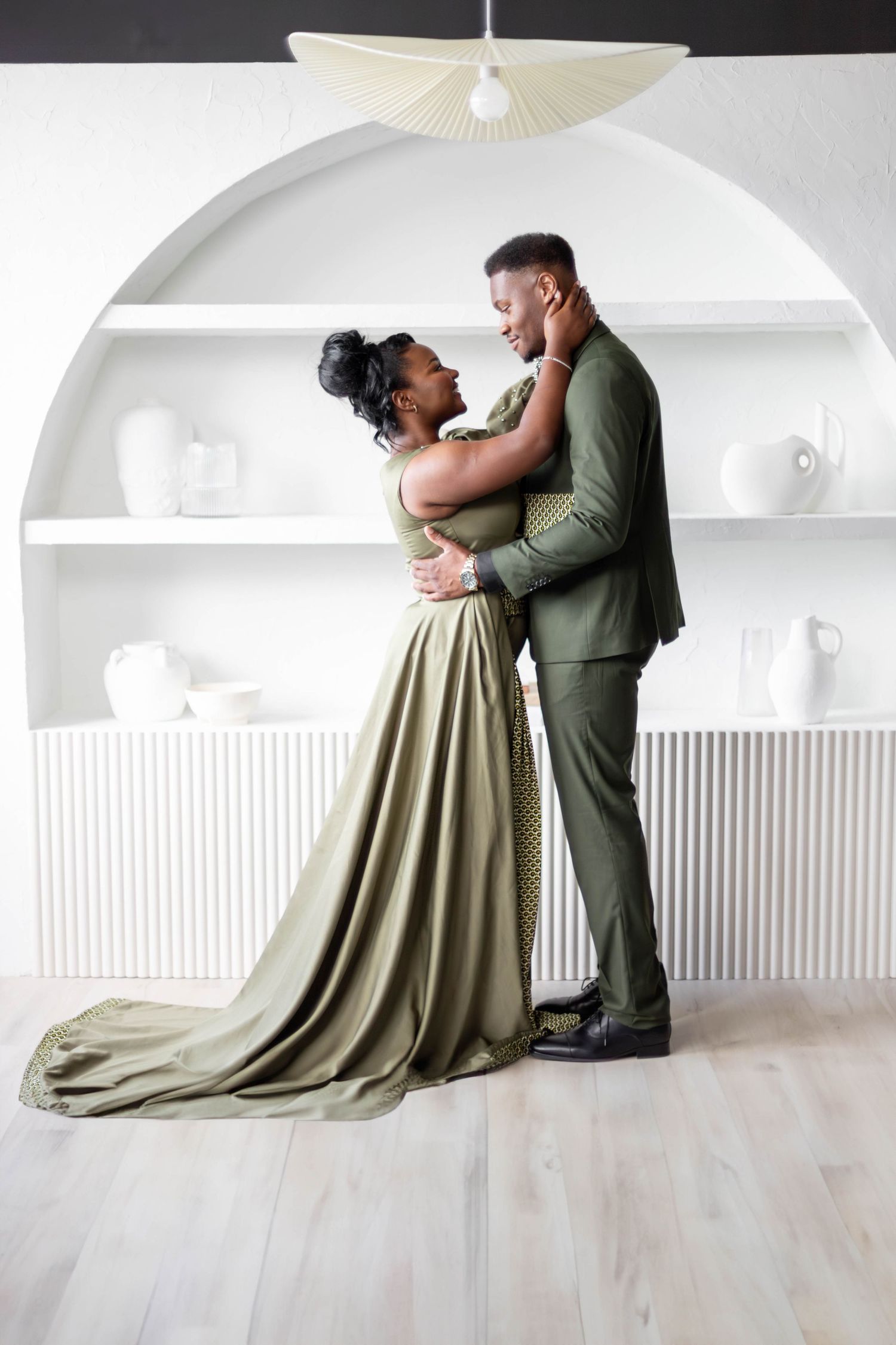 A couple embraces in elegant olive green formal wear against a white backdrop.