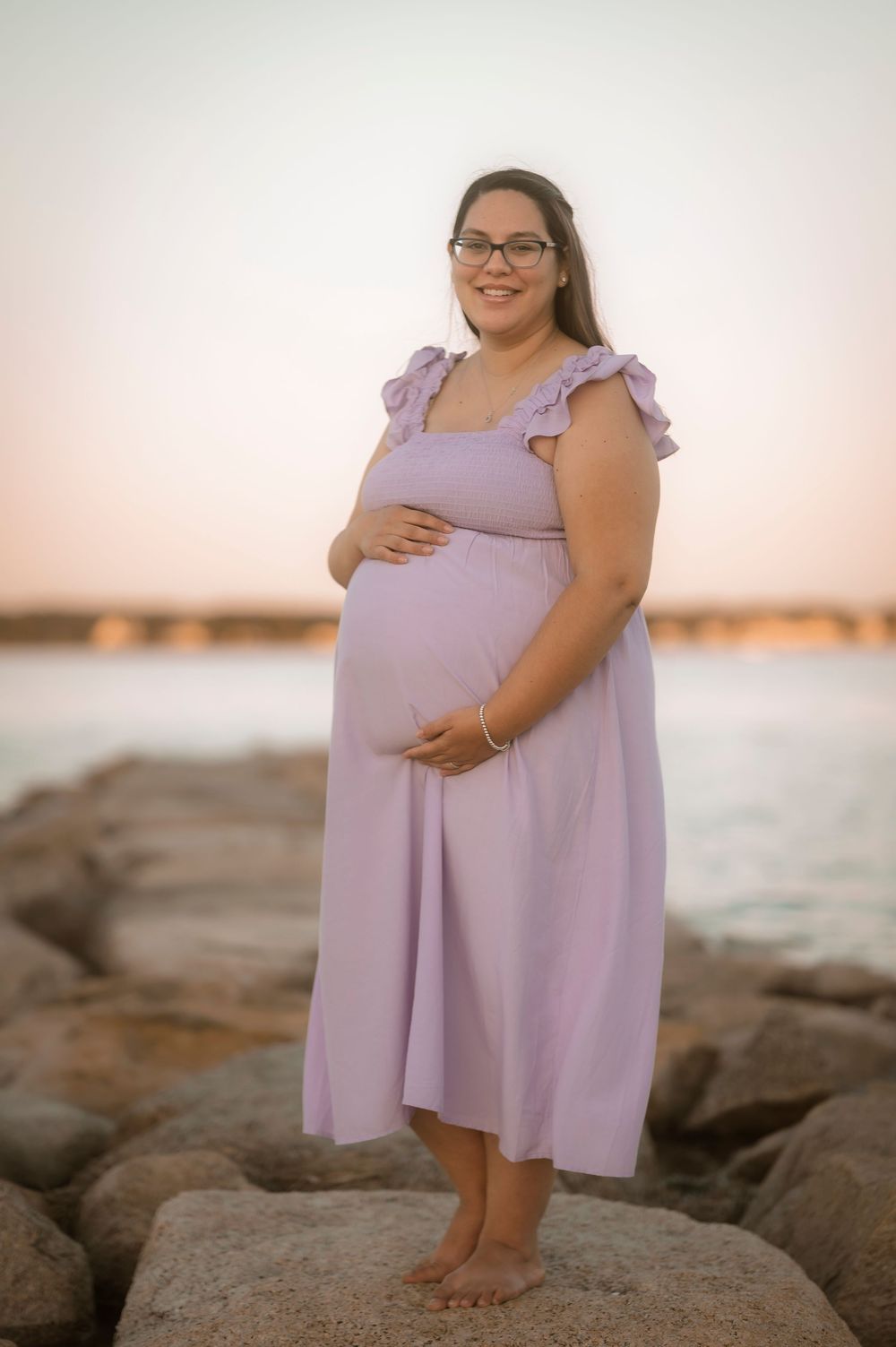 A pregnant woman in a lavender dress poses on coastal rocks at sunset.