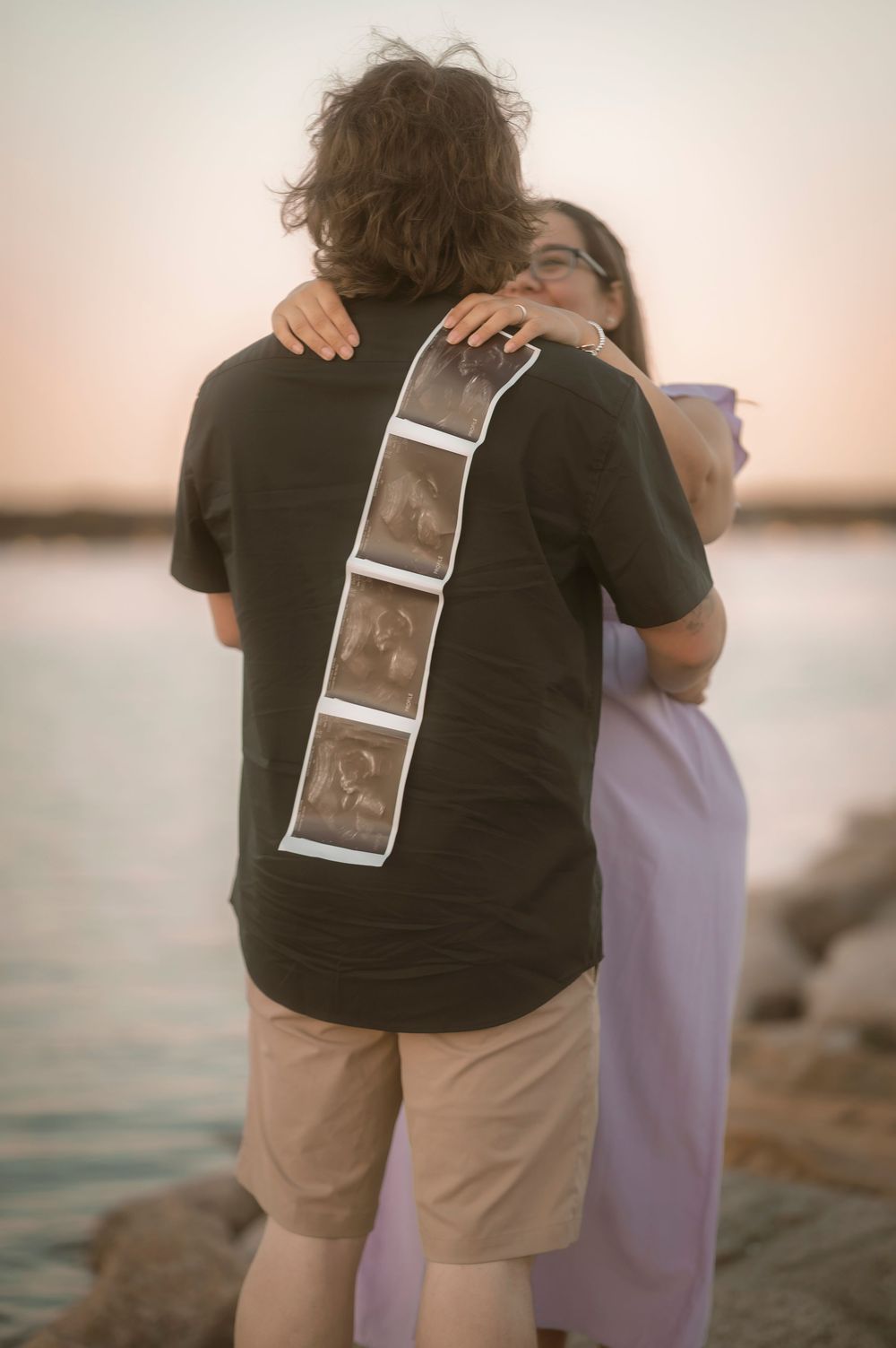 A couple embracing on a rocky shore at sunset while holding ultrasound photos together against the water.