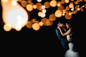 Romantic wedding couple embracing under warm glowing bokeh string lights in dark reception venue. Fotógrafo de Bodas en Formosa