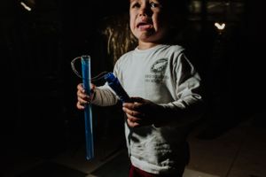 Young child holds a blue tube and toy in a dimly lit room, wearing a white shirt and red pants. Fotógrafo de Bodas en Formosa