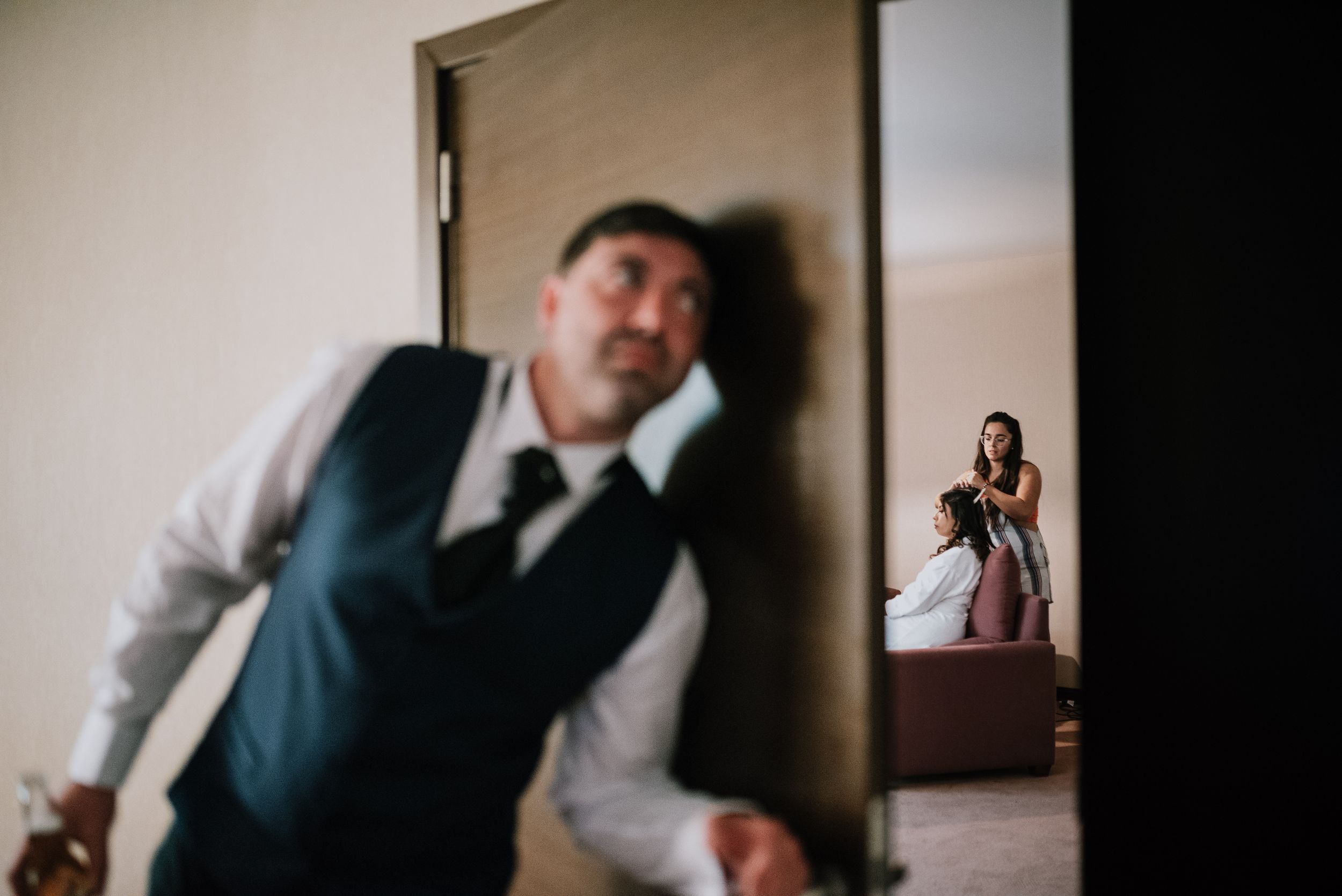 Groom leaning against door frame while bride photographs him in mirror reflection during wedding preparations. Fotógrafo de Bodas en Formosa