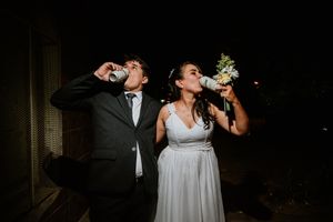 Bride and groom drink from cans outdoors at night, bride holding a floral bouquet in a white gown. Fotógrafo de Bodas en Formosa